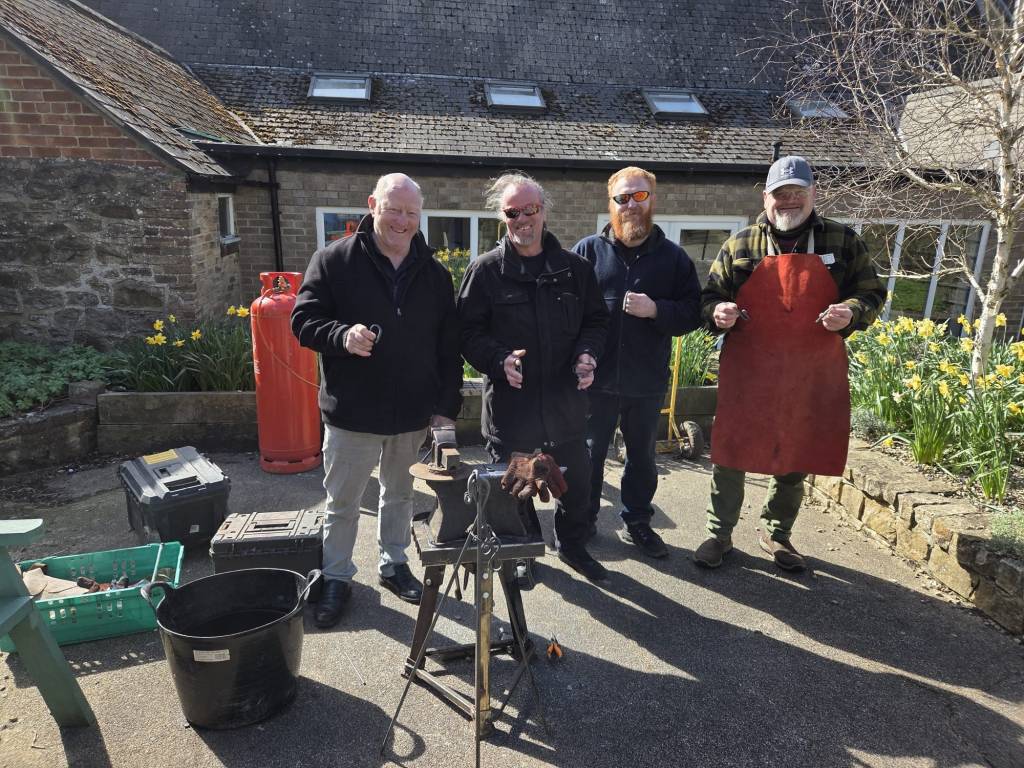 A group of people who have enjoyed a blacksmith course, smiling stood around an anvil